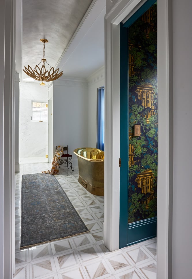 A beautifully designed bathroom with a large copper bathtub, a copper chandelier, a small iron chair, and a long gray rug on geometric flooring. The room has a classic domed ceiling, marble walls, and bright white accents on the crown molding. 