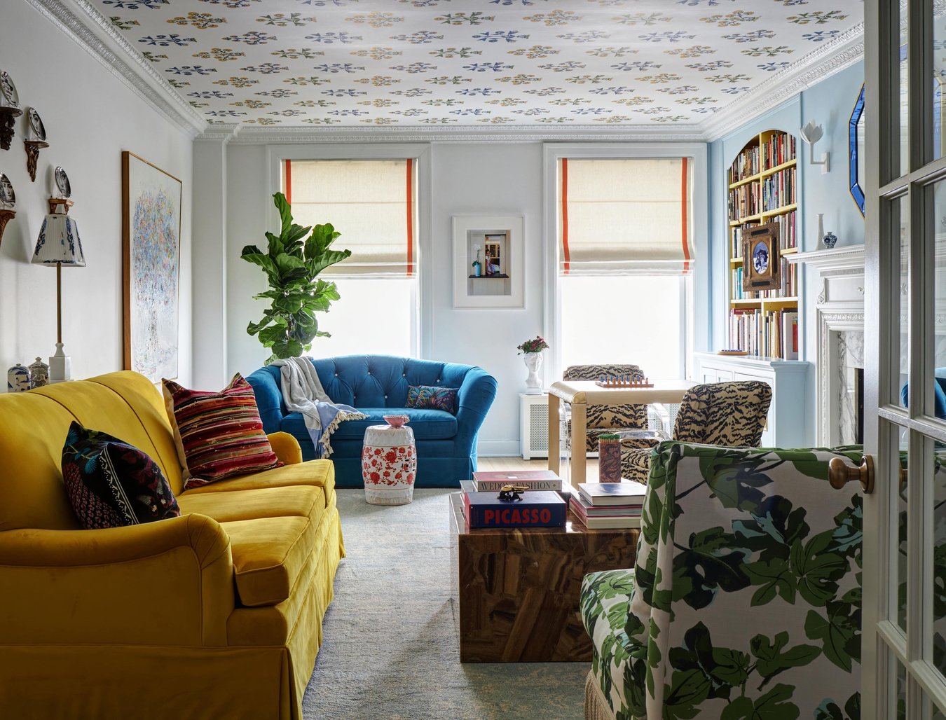 A bright living room with a bright yellow sofa and a bright blue loveseat is accented by large print art, inset bookshelves, a marble and white fireplace, a lush fig plant, and a table with tiger print chairs, all beneath a floral print ceiling - living room design by Jasmin Reese.