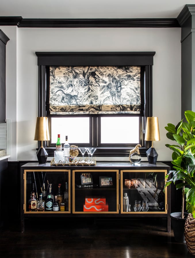 An antique, dark wood, glass, and brass bar with black and gold lamps against a window with a black and cream jungle print shade - living room design by Jasmin Reese Interiors, Chicago, USA. 