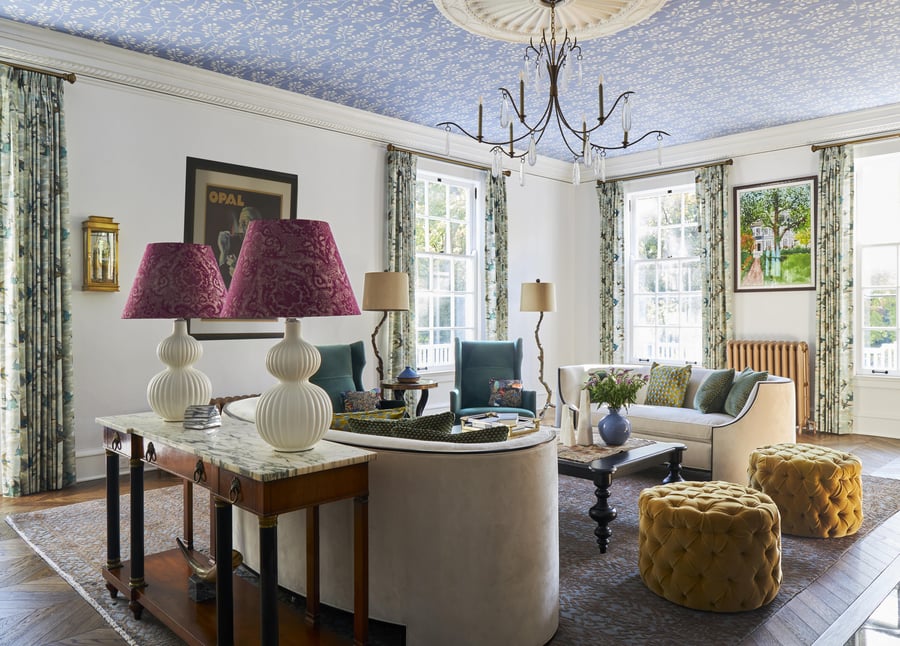 A large living room with double cream rounded sofas with puff ottoman in gold with an antique marble and wood accent table with two white lamps with red paisley lampshades beneath a periwinkle and white floral ceiling - living room design by Jasmin Reese Interiors, Chicago, USA. 