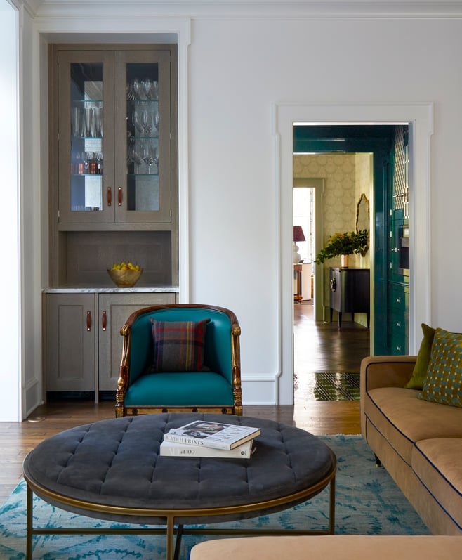 A turquoise leather armchair behind a gray metal and velvet ottoman with a dry bar in the background - living room design by Jasmin Reese Interiors, Chicago, USA. 