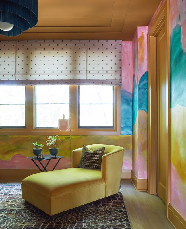 A bright yellow plush chaise lounge in a roomy corner of a room painted in sunset colors of pink, yellow, orange, green, and blue beside a window with a white and navy polka dot drop shade - living room design detail by Jasmin Reese Interiors, Chicago, USA. 