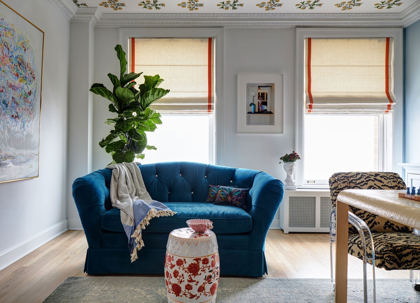 A bright blue plush loveseat in a small apartment with a small antique coffee table in red floral prints - the room is full of natural light, art, and plants - living room design by Jasmin Reese Interiors, Chicago, USA. 
