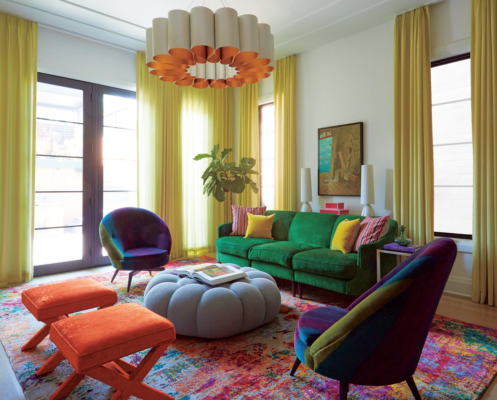 A colorful living room with a plush green sofa, light blue puff ottoman coffee table, two plush orange stools, and a blue, green, and purple velvet rounded chair with natural light streaming through lime green curtains. An orange and cream modern enamel chandelier centers the room over a multicolored area rug - living room design by Jasmin Reese Interiors, Chicago, USA. 