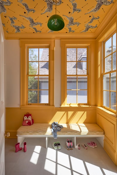 A bright yellow and white mudroom with an upholstered bench beneath tall windows. The bench has children's shoes beneath and a backpack and jacket - interior design by Jasmin Reese Interiors, Chicago, USA. 