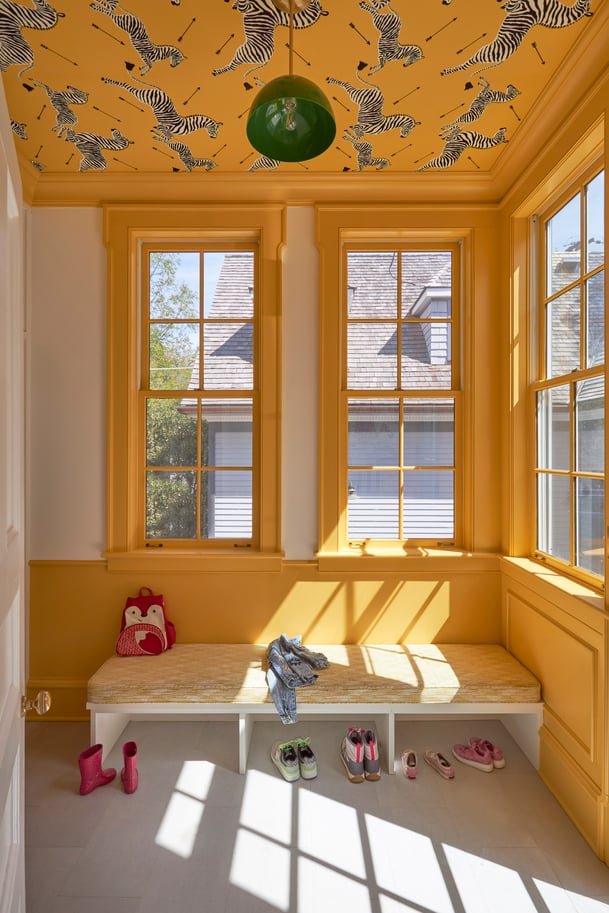 A bright yellow and white mudroom with an upholstered bench beneath tall windows. The bench has children's shoes beneath and a backpack and jacket - interior design by Jasmin Reese Interiors, Chicago, USA. 