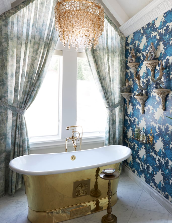  A side view of a large marble bathroom with a brass bathtub and chandelier designed by Jasmin Reese, Chicago. 