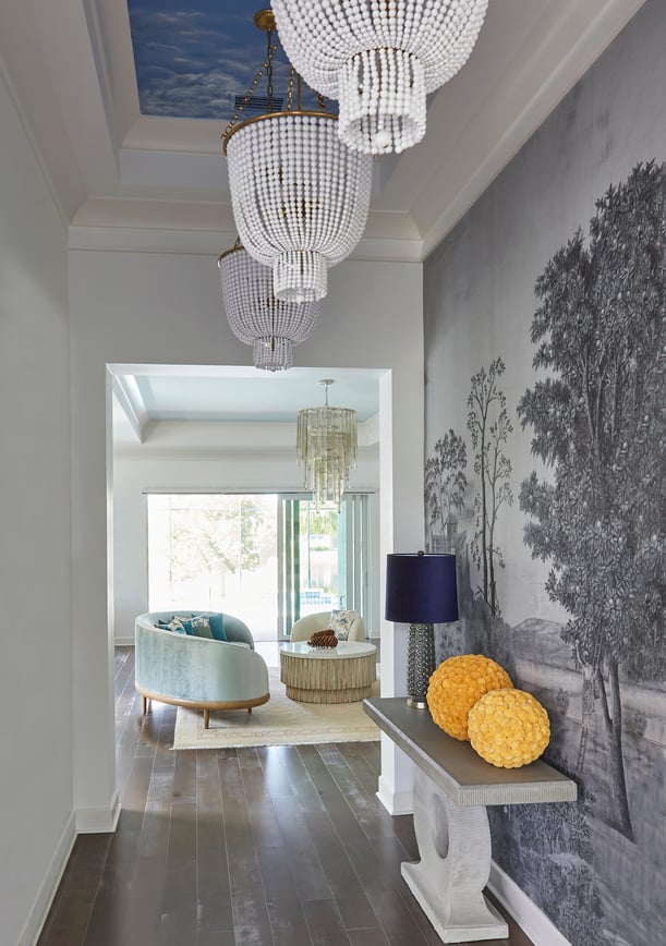 A view of a seafoam green circular sofa in a living room seen from a hallway with bright blue lamps and wallpaper featuring pastoral trees in gray - living room design by Jasmin Reese Interiors, Chicago, USA. 