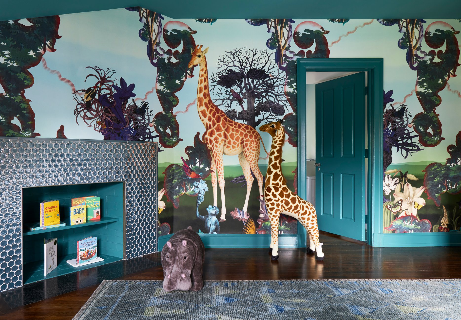 A child's playroom with a stuffed giraffe and hippo next to an old fireplace turned into a bookshelf. The room has wallpaper with giraffes, monkeys, trees, tropical flowers, and woodpeckers - interior design by Jasmin Reese Interiors, Chicago, USA. 