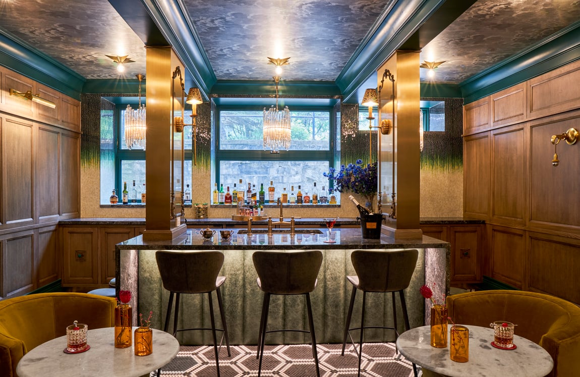 A warm, dreamy bar area with a marble bar with under-the-table lighting with three low-back barstools, crystal chandeliers, geometric woven rugs, round marble-top tables with gold velvet circular armchairs in the forefront - interior design by Jasmin Reese Interiors, Chicago, USA. 