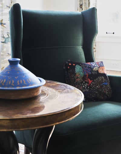 A plush forest green wingback chair next to a high round wooden side table with a blue and gold rustic vase - living room design detail by Jasmin Reese Interiors, Chicago, USA. 