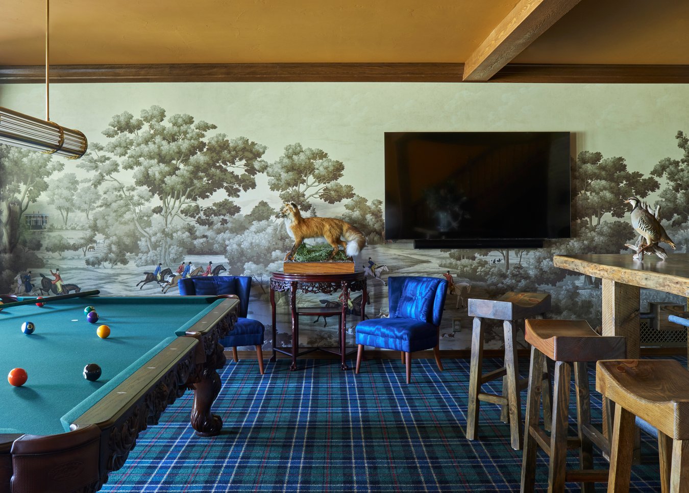 A rustic den with a pool table, flat-screen TV, a rustic wood bar with barstools, bright blue chairs, flannel carpet, taxidermy fox, and wallpaper depicting a fox hunt through the trees - interior design by Jasmin Reese Interiors, Chicago, USA. 