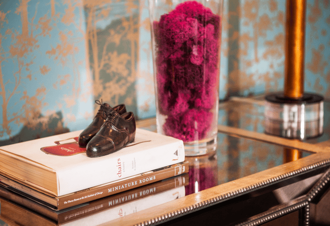 A miniature pair of wingtip leather shoes sitting atop two books about miniatures in design next to a large glass vase full of magenta roses against turquoise and gold foil wallpaper - living room design by Jasmin Reese Interiors, Chicago, USA. 
