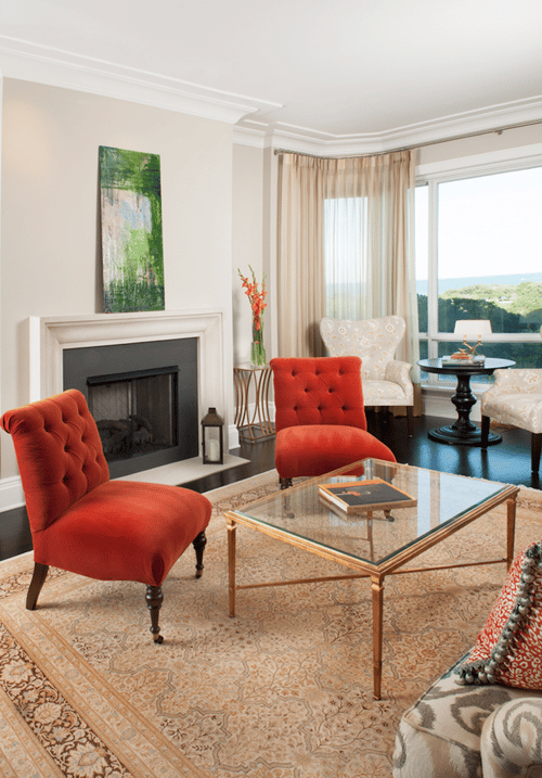 Two orange velvet clutch chairs around a glass and brass coffee table in front of a black fireplace with a low white mantle with an abstract painting above it - living room design by Jasmin Reese Interiors, Chicago, USA. 