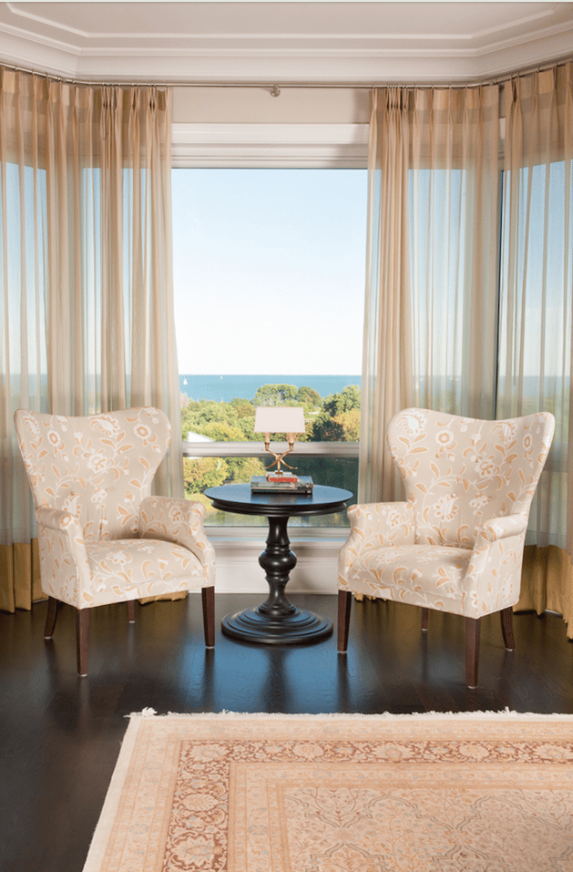Two cream and tan wingback chairs with a floral print in tan around a dark wood circular table with a small brass lamp in front of a large window with transparent tan drapes - living room design by Jasmin Reese Interiors, Chicago, USA. 
