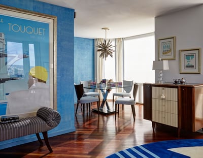 A view of a small dining area with bright turquoise walls and a large window with four modern chairs around a glass table covered in colorful books - living room design by Jasmin Reese Interiors, Chicago, USA. 