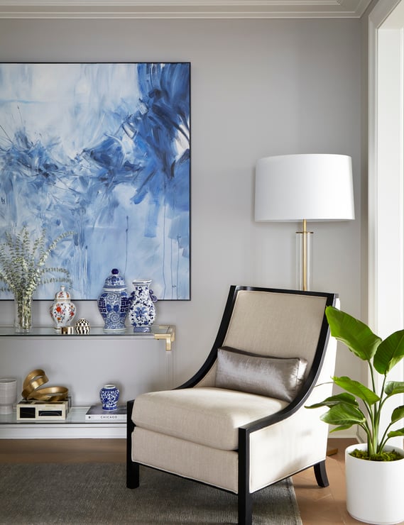 A wing-back chair with dark wood and cream upholstery beside a long glass table beneath a large impressionist painting in blues and white oil - living room design by Jasmin Reese Interiors, Chicago, USA. 