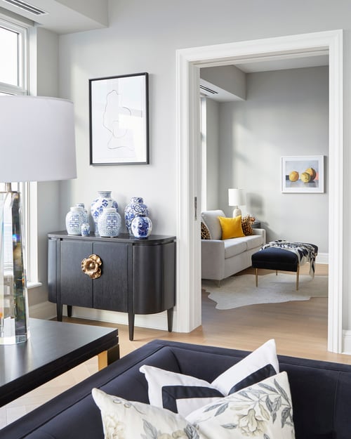 A view of a small white living room sofa and a blue ottoman off the main living room with a large blue sofa and an antique sideboard with antique vases - living room design by Jasmin Reese Interiors, Chicago, USA.