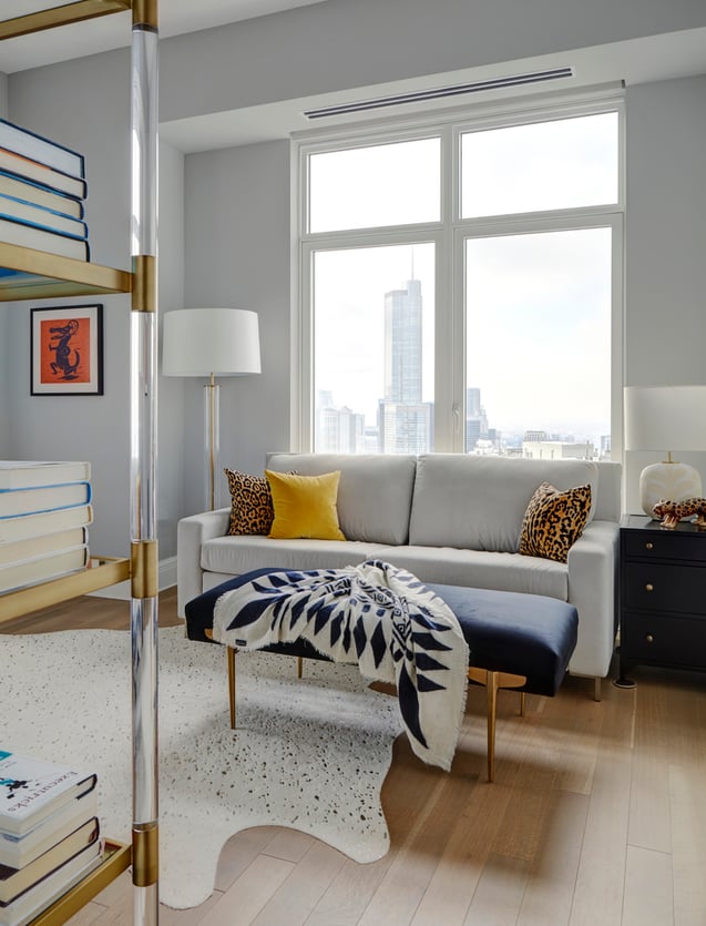 A small white sofa in front of a large cityscape window with a dark blue ottoman and reading lamps. A glass and gold bookshelf sits in the forefront - living room design by Jasmin Reese Interiors, Chicago, USA.