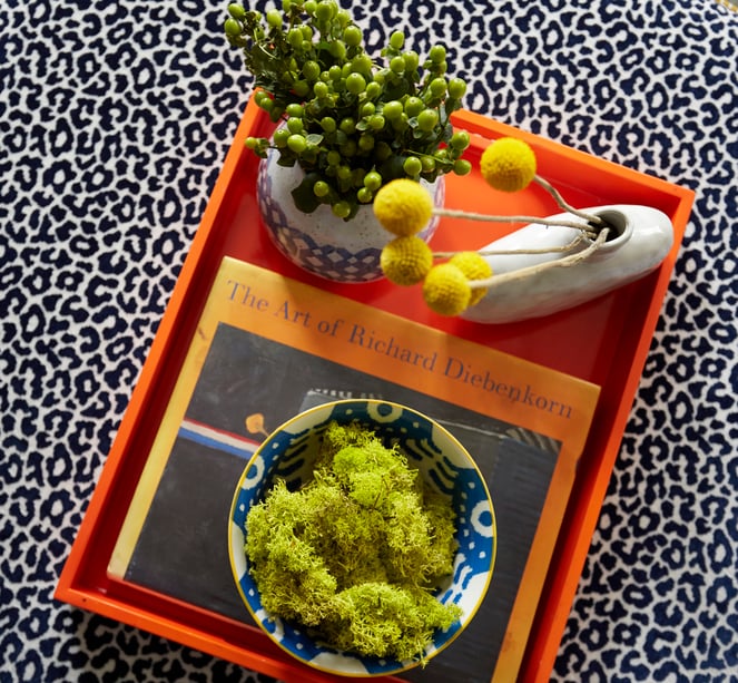 An orange tray on a leopard print fabric ottoman in a living room designed by Jasmin Reese: neon green moss, berries, and tuft flowers in vases. 