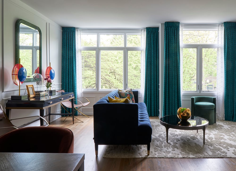 A long blue sofa with a black enamel and wood coffee table and a small circular green clutch chair against tall windows with textured green drapes - living room design by Jasmin Reese Interiors, Chicago, USA.