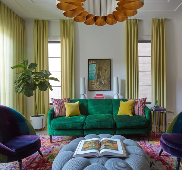 A plush green sofa and a light blue puff ottoman coffee table with natural light streaming through chartreuse curtains. An orange and cream modern enamel chandelier centers the room over a multicolored area rug - living room design by Jasmin Reese Interiors, Chicago, USA. 