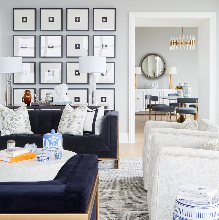 A view through a living room with a long dark grey sofa with a long table with glass lamps in front of a collection of sixteen framed pictures of a black square with a white circle inside at another room with a small dining table - living room design by Jasmin Reese Interiors, Chicago, USA.