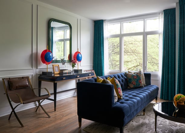 A long blue sofa with a black enamel and wood coffee table with an antique leather camp chair beside a black enamel side table with bright red and blue art pieces around a rounded square mirror - living room design by Jasmin Reese Interiors, Chicago, USA.