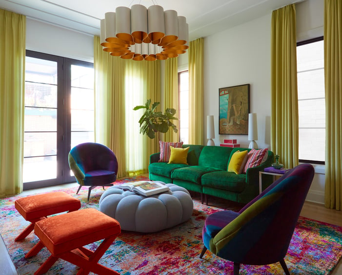 A colorful living room with a plush green sofa, light blue puff ottoman coffee table, two plush orange stools, and a blue, green, and purple velvet rounded chair with natural light streaming through chartreuse curtains. An orange and cream modern enamel chandelier centers the room over a multicolored area rug - living room design by Jasmin Reese Interiors, Chicago, USA. 
