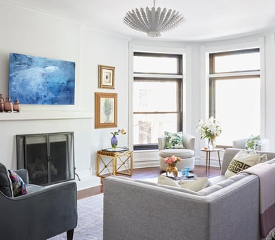 A bright white living room with a tan sofa and a dark grey armchair in front of a minimalist fireplace with an abstract blue painting above -  living room design by Jasmin Reese Interiors, Chicago, USA.