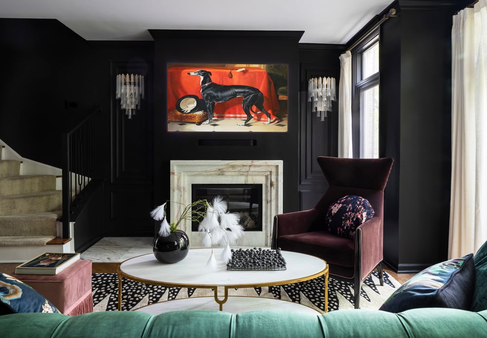 A warm living room painted black with a modernist marble fireplace, art deco sconces, a large brown velvet wing-back chair, and an oval marble and brass coffee table with a painting of a greyhound over the fireplace - living room design by Jasmin Reese Interiors, Chicago, USA. 