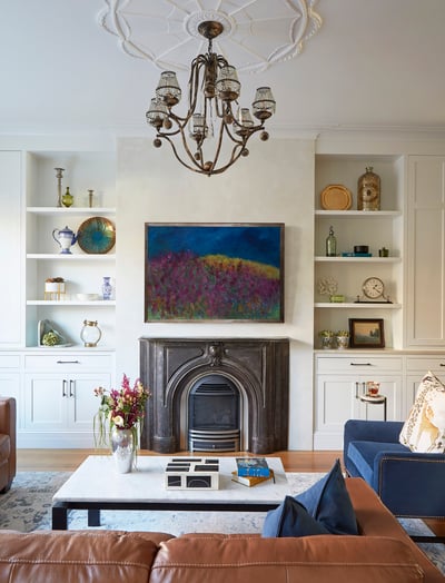 A bright white living room with an antique metal fireplace, brown leather sofa, and built-in bookshelves around a large art piece of a field of bright magenta flowers - living room design by Jasmin Reese Interiors, Chicago, USA.