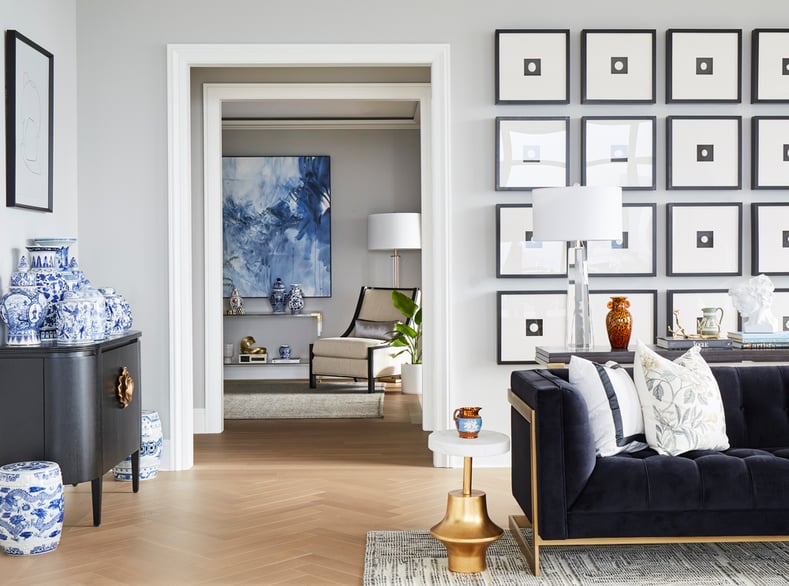  A view through a living room with a long dark grey sofa with a long table with glass lamps in front of a collection of sixteen framed pictures of a black square with a white circle inside at another room with a white chair and a large blue painting - living room design by Jasmin Reese Interiors, Chicago, USA.