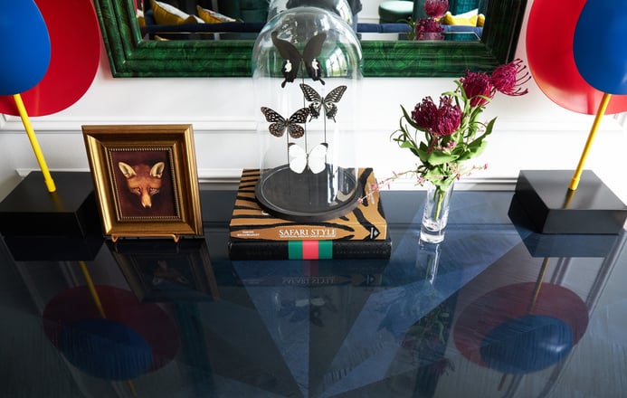 A detail of four butterflies under a glass dome on two books about safari beside a painting of a fox in a gold frame on a black enamel tabletop - living room design by Jasmin Reese Interiors, Chicago, USA. 