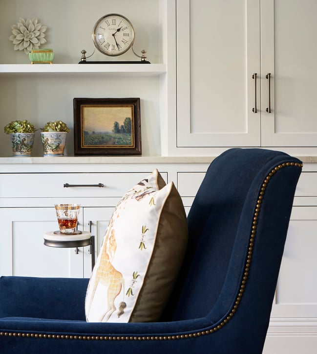 A side view of a blue velvet armchair beside a built-in bookshelf with an antique tabletop clock on a top shelf - living room design by Jasmin Reese Interiors, Chicago, USA. 