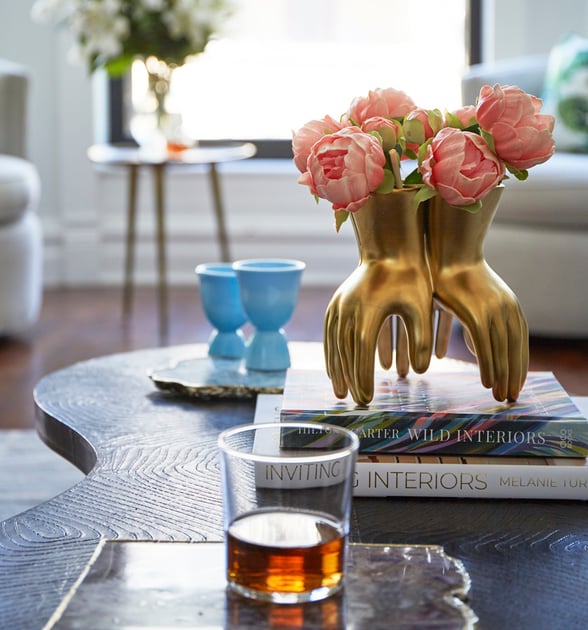 A gold vase in the shape of three hands with pink peonies atop two books on a wooden coffee table with a bourbon glass - living room design by Jasmin Reese Interiors, Chicago, USA.