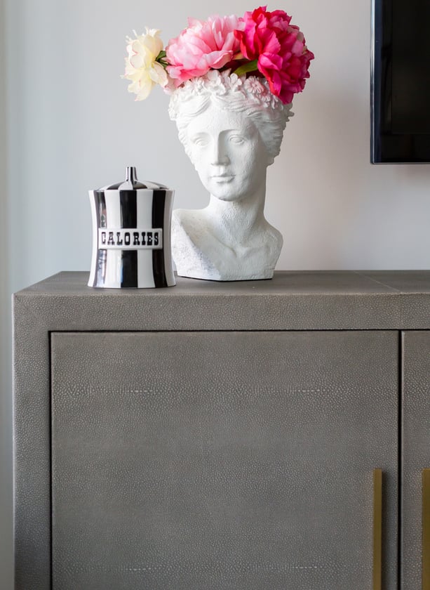 A textured grey leather chest with a white vase in the shape of a goddess with bright pink peonies with a black and white striped ceramic jar with the word 