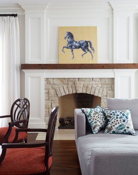 A painting of a horse in blue and gold over a rough white stone fireplace beside a grey chaise lounge and two armchairs with red cushions - living room design by Jasmin Reese Interiors, Chicago, USA. 